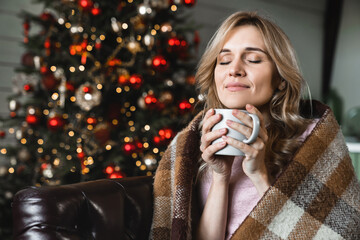 Caucasian woman wrapped warm blanket relaxing at home, sitting on cozy couch, happy smiling female drinking tea or coffee, dreaming, enjoying winter holidays with Christmas tree in the background