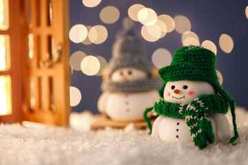 Cute decorative snowmen and lantern on artificial snow against blurred lights, selective focus