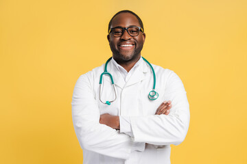 Happy African American man wearing doctor uniform with crossed arms isolated on yellow