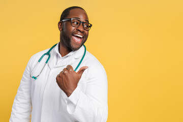 Happy African American doctor in glasses pointing to copy space by thumbs up in studio