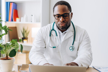 Confident African American doctor in eyeglasses working with laptop typing