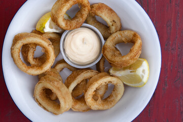 Delicious fried seafood. Closeup view of fried squid rings with lemon and a dipping sauce in a white bowl on the wooden table.