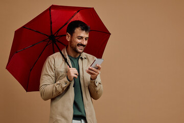 Happy man under red umbrella texting on cell phone.