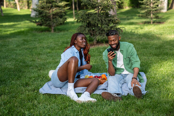 Joyful African American couple enjoying relaxed picnic in park, sitting on grass and chatting happily. Pleased black students savor quality time together, sharing good mood with smiles outdoor setting