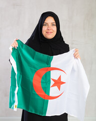 smiling Muslim woman in traditional black hijab holds flag of Algeria. Portrait of female Muslim...