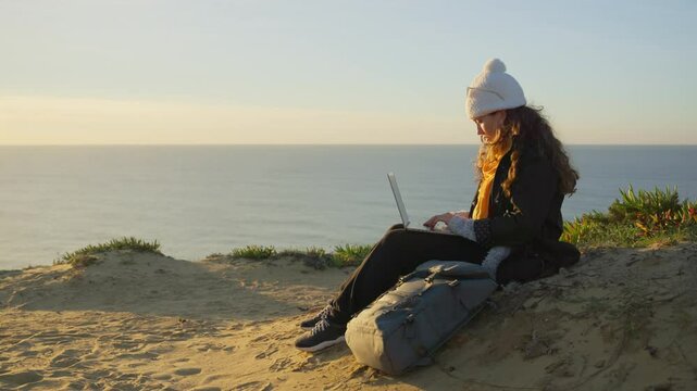 Female typing on laptop sitting on cliff by the ocean enjoying working outside. Woman freelancer or student digital nomad is using computer outdoors in nature with beautiful work environment 