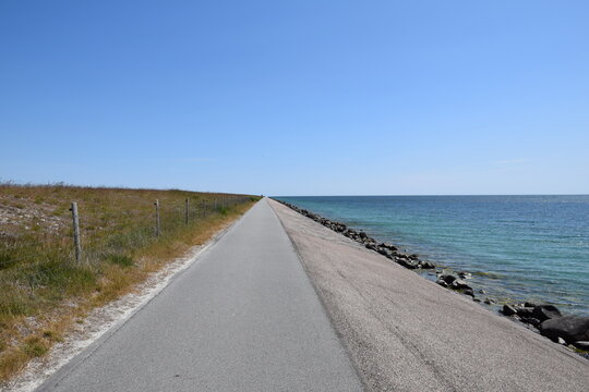 View of paved bicycle road extending towards horizon, summer vegetation on the left, sloping concrete, rocks and blue ocean on the right, blue skies, summer time, August, Amager Island, Denmark