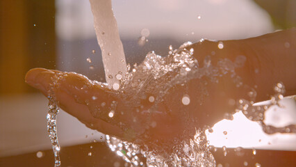 Fototapeta premium CLOSE UP, LENS FLARE: Crystal clear water splashes over an open hand beneath a stainless steel kitchen faucet, with morning sunlight illuminating each droplet. Fresh running water in modern household.