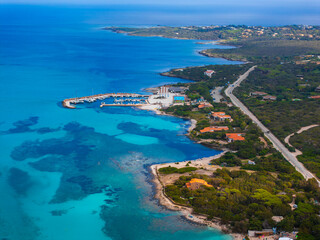 Aerial view of Sardinia, Italy, showcasing a turquoise sea, a small marina with boats, lush greenery, and scattered buildings along a winding road.