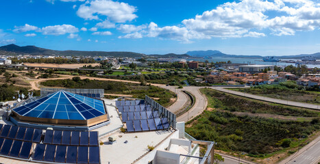 Aerial view of Olbia, Sardinia, Italy, featuring a modern building with a glass dome and solar panels. The scene includes urban areas, green fields, and a harbor.