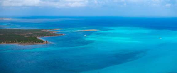 Aerial view of Sardinia's coastline with turquoise waters, a solitary tower, and scattered boats. The partly cloudy sky adds depth to the scene.