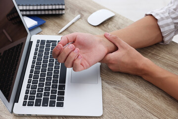 Man suffering from pain in wrist while working on laptop at table indoors, closeup. Carpal tunnel...