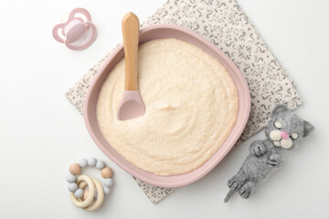 Delicious baby food in bowl with spoon, pacifier and toys on white background, flat lay