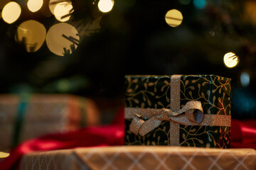 Close up background image of tiny Christmas present with golden ribbon hiding under Christmas tree with blurred twinkling lights copy space