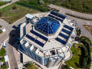 An aerial view captures a modern octagonal building with a blue glass dome, surrounded by solar panels and greenery, located in Sardinia, Italy.