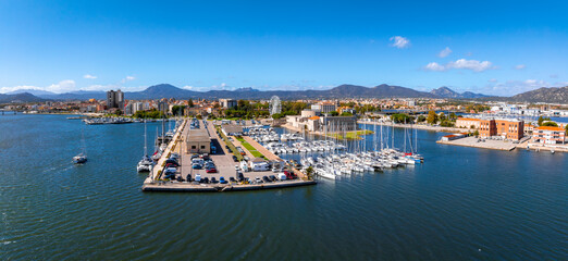 Aerial view of Olbia, Sardinia, featuring a marina with yachts, a Ferris wheel, and surrounding mountains under a clear blue sky and calm waters.