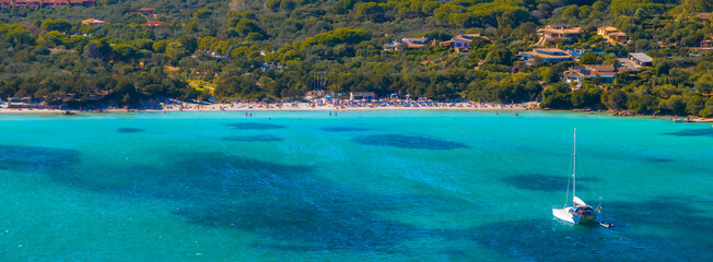 Aerial view of a Sardinian beach with turquoise waters, a sailboat, sunbathers, umbrellas, and villas nestled in lush greenery.