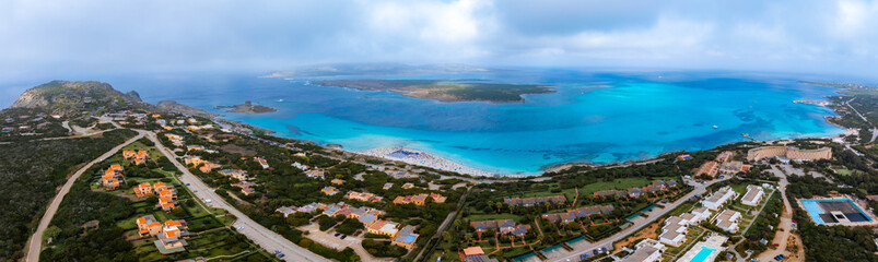 Aerial view of Sardinia, Italy, showing turquoise Mediterranean waters, sandy beaches, and orange roofed houses nestled in lush greenery.