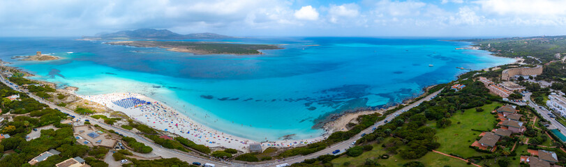 Aerial view of Torre della Pelosa on Sardinia's coast, featuring turquoise waters, white sand, colorful umbrellas, and lush greenery in Italy.