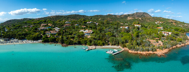 Naklejka premium Aerial view of a Sardinian beach with turquoise waters, sunbathers, and boats. Hillside villas and lush greenery enhance the Mediterranean landscape.
