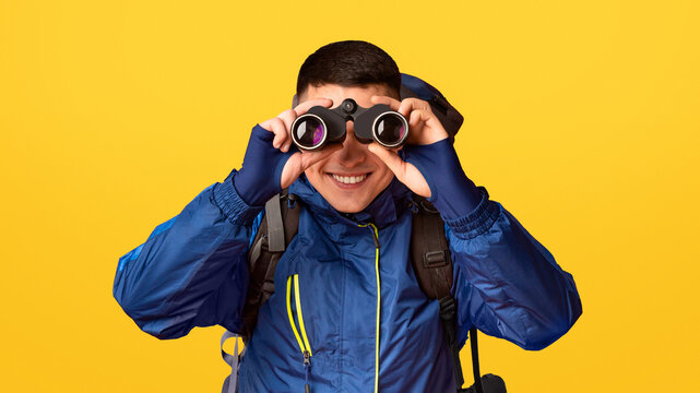 Portrait of smiling young guy hicker using modern binoculars, orange background