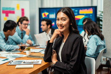 Portrait of happy young asian businesswoman or analyst looking at camera with her colleague analyzing data analysis in dynamic business strategy investment planning meeting. Habiliment
