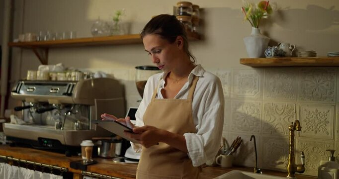 A confident girl pastry chef in a white shirt and a beige apron keeps track of goods using an electronic tablet in the kitchen of a bakery