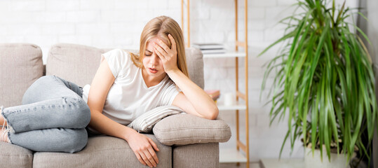 Upset young woman laying on couch at home, suffering from headache, panorama with copy space