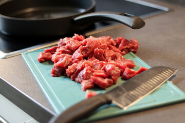 Close up of raw beef meat chopped in small cubes on green cutting board with a knife
