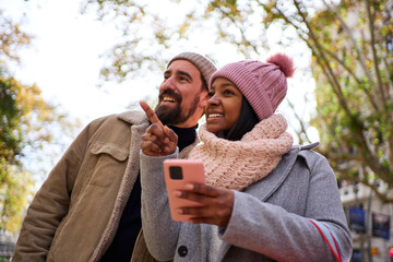 Front view of a young multiethnic couple using phone together to look for a direction during winter holidays in the city, pointing at something.