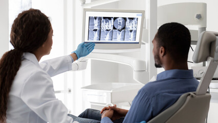 Female dentist african woman pointing at screen with xray picture, showing patient his teeth