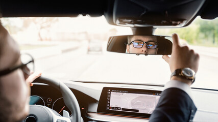 Close up back view portrait of man in specs adjusting rearview mirror while sitting in his car, selective focus