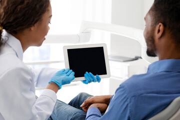 Close up of african female dentist and male patient looking at empty tablet screen, technologies in dentistry