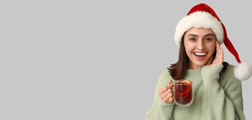 Happy young woman in Santa hat holding glass cup of hot mulled wine on grey background with space for text