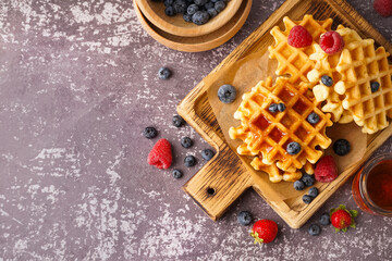 Wooden board of tasty waffles with berries and maple syrup on grey background