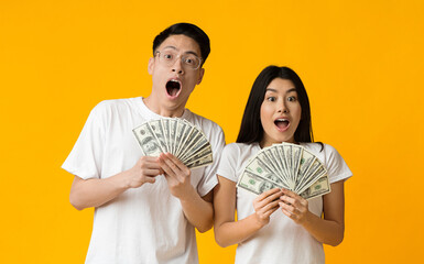 Amazed asian couple holding bunch of money banknotes and celebrating, yellow studio background