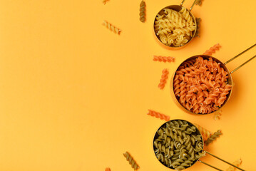 Metal scoops with different types of delicious uncooked pasta on orange background