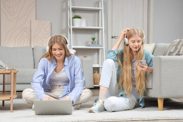 Beautiful young happy sisters in headphones with laptop and mobile phone sitting on floor at home