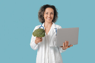 Mature female nutritionist with broccoli and laptop on blue background