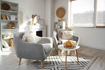 Interior of light living room with grey armchairs, fireplace, pumpkins and white shelving unit