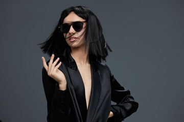 A stylish woman in a black suit and sunglasses poses confidently for the camera with her hand on her hip and a cigarette in her mouth, exuding a sense of empowerment and cool sophistication