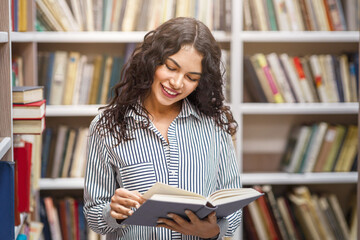 Learning Concept. Smiling mexican girl standing at bookshelves in campus library, reading book,...