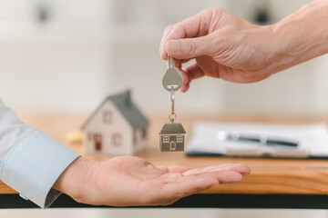 A hand passes house keys to another, symbolizing the transfer of property ownership, with a miniature house in the background.