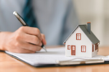 A person writes on a document next to a miniature house, symbolizing real estate transactions or...