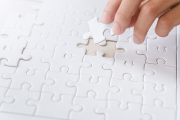 A hand is placing a missing white puzzle piece into a nearly complete puzzle on a flat surface.