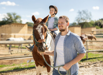 Young man leads horse on leash with boy rider in paddock