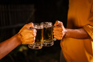 A man's hand and a woman's hand hold beer glasses in a cozy, lively evening party setting. Family and friends gather under the night sky, sharing joyful moments and laughter.