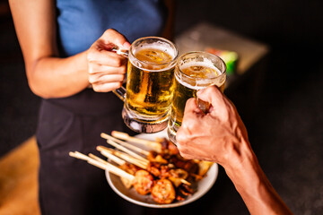 A man's hand and a woman's hand hold beer glasses in a cozy, lively evening party setting. Family and friends gather under the night sky, sharing joyful moments and laughter.