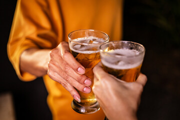 A man's hand and a woman's hand hold beer glasses in a cozy, lively evening party setting. Family and friends gather under the night sky, sharing joyful moments and laughter.
