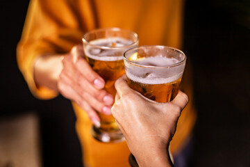 A man's hand and a woman's hand hold beer glasses in a cozy, lively evening party setting. Family and friends gather under the night sky, sharing joyful moments and laughter.